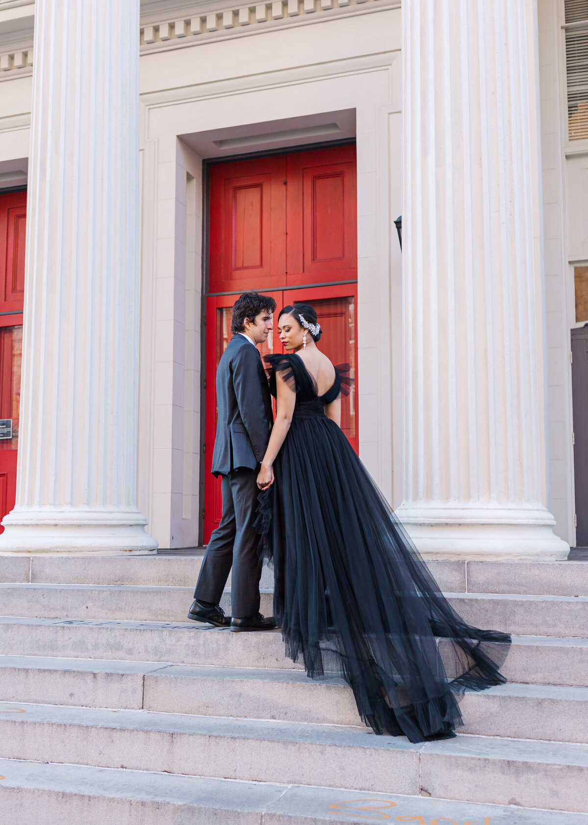 woman wearing black tulle dress and man wearing dark gray suit holding hands on marble steps in front of red door