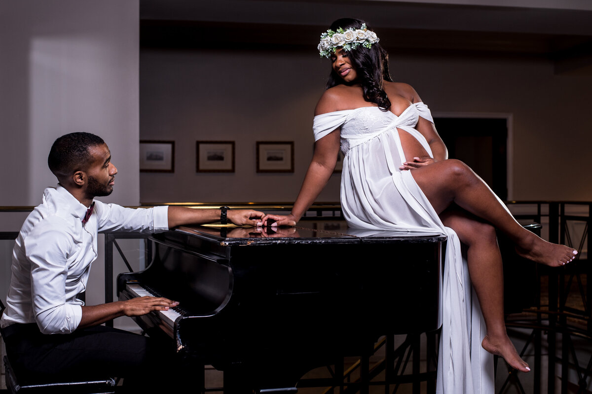 a couple sit on and next to a black piano. The woman wears a white maternity dress and a flower crown