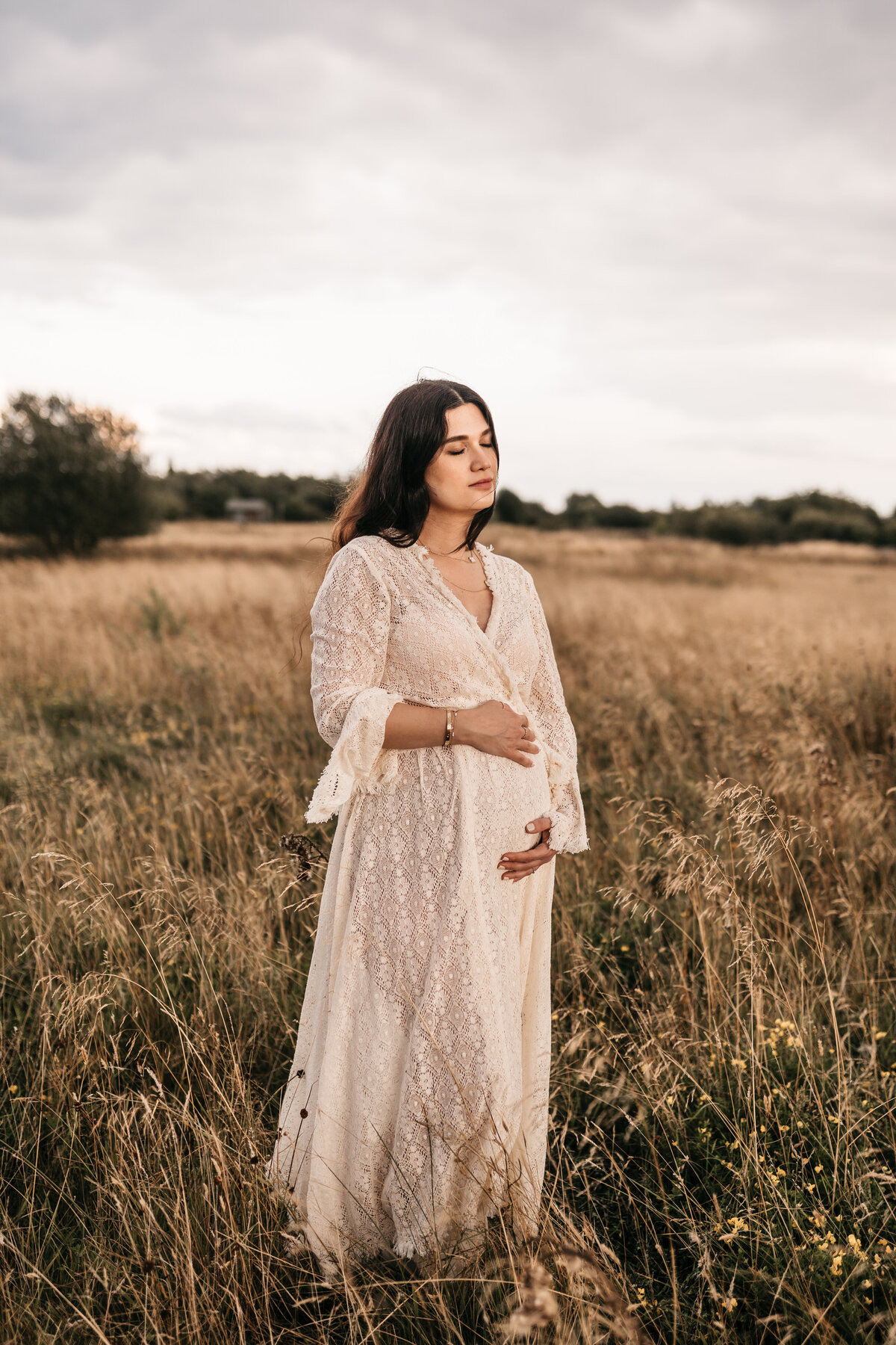 Photo of a pregnant woman standing in a field holding her bump with her eyes closed