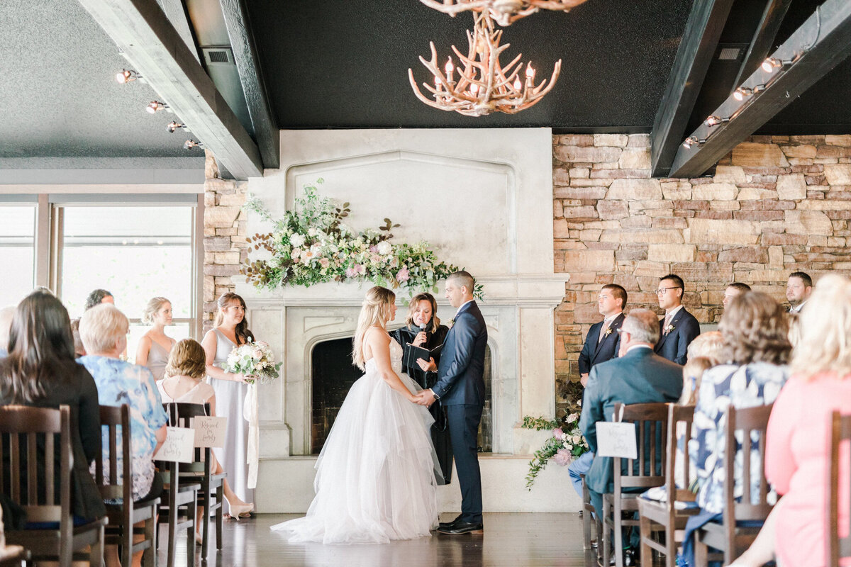 A wedding ceremony at The Lake House in Calgary, featuring a couple exchanging vows in front of a stone fireplace adorned with lush floral arrangements. The bride wears a flowing white gown, and the groom is in a navy-blue suit. Bridesmaids in soft pastel dresses stand to the left, holding bouquets, while groomsmen in matching suits stand to the right. Guests are seated on wooden chairs, attentively watching the moment. The venue’s rustic charm is highlighted by exposed wooden beams, an antler chandelier, and large windows allowing natural light to fill the space.
