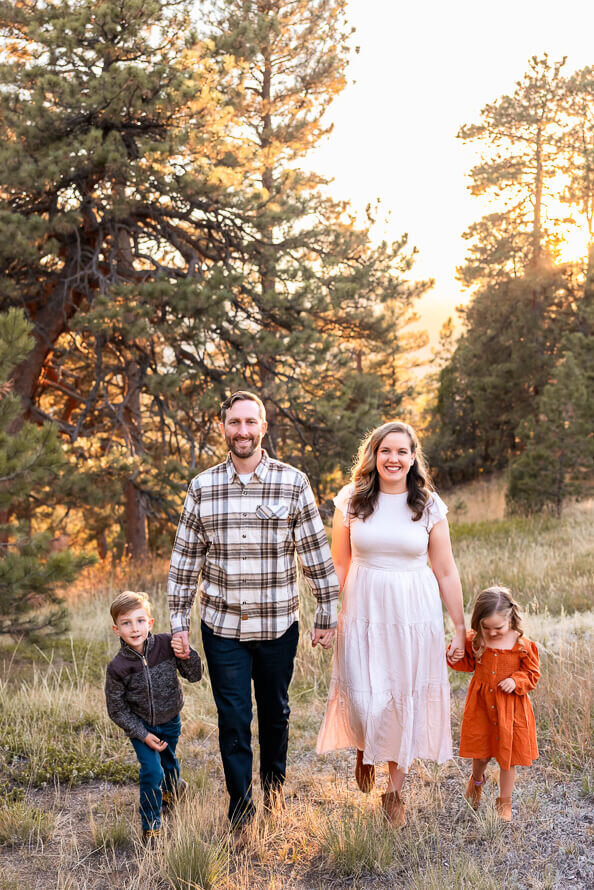 Family holds hands walking through the forest with golden sunlight behind them.
