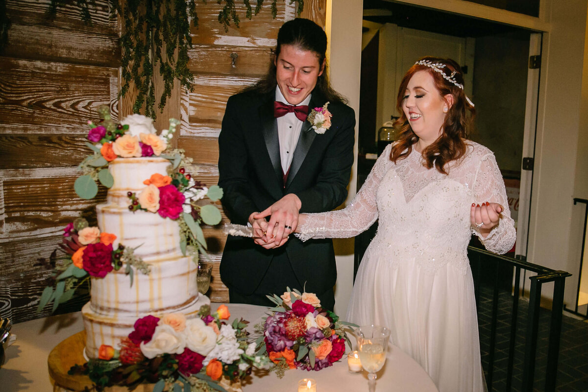 A bride and groom, smiling, cut their wedding cake together. The cake is decorated with colorful flowers and sits on a table with more floral arrangements and candles. The couple looks happy and excited.