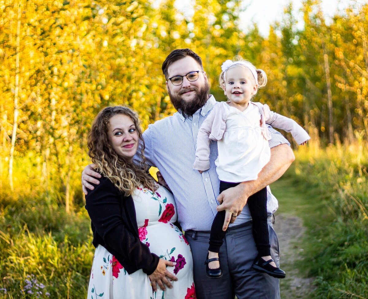 A woman in a floral dress holds her pregnant belly next to her husband who is holding their toddler daughter
