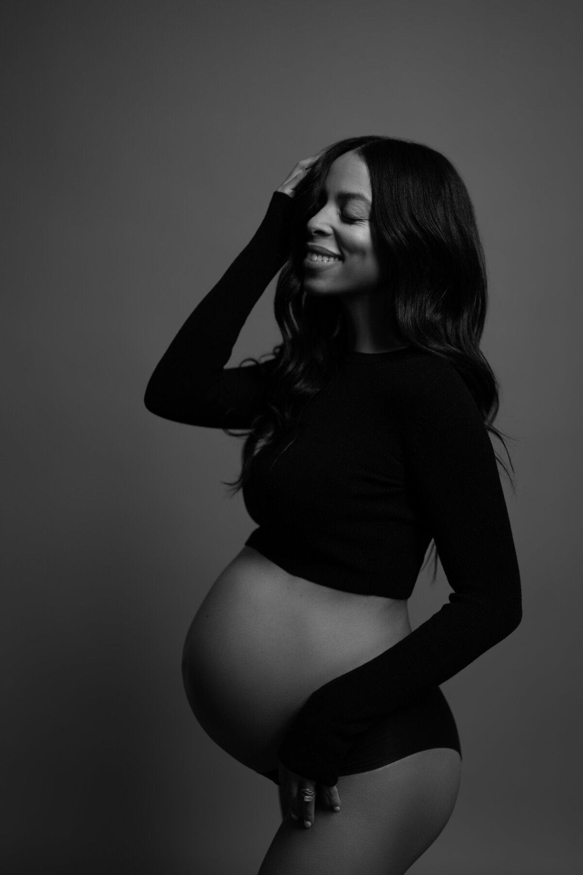 A striking black-and-white maternity portrait featuring the expectant mother posing gracefully with her hands overhead, taken in a professional studio in Brooklyn, NY.