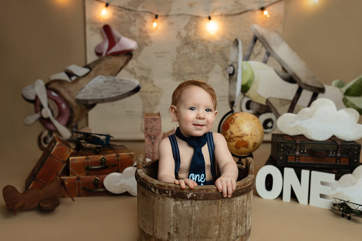 A toddler boy sits in a wooden bucket in suspenders and a tie