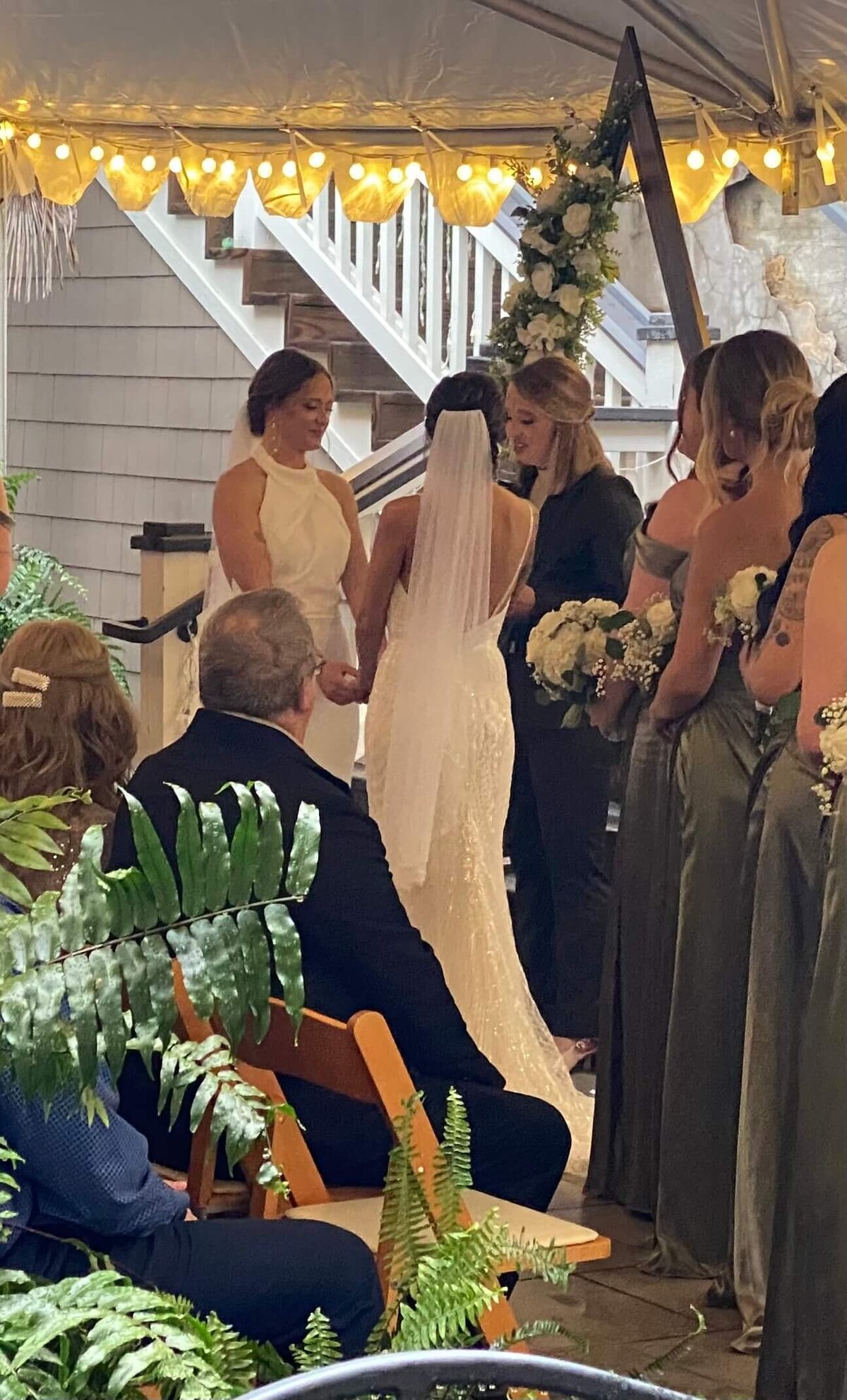 Two brides stand holding hands during their wedding ceremony under a tent, surrounded by bridesmaids in green dresses and guests seated on wooden chairs. The scene is decorated with greenery and string lights.