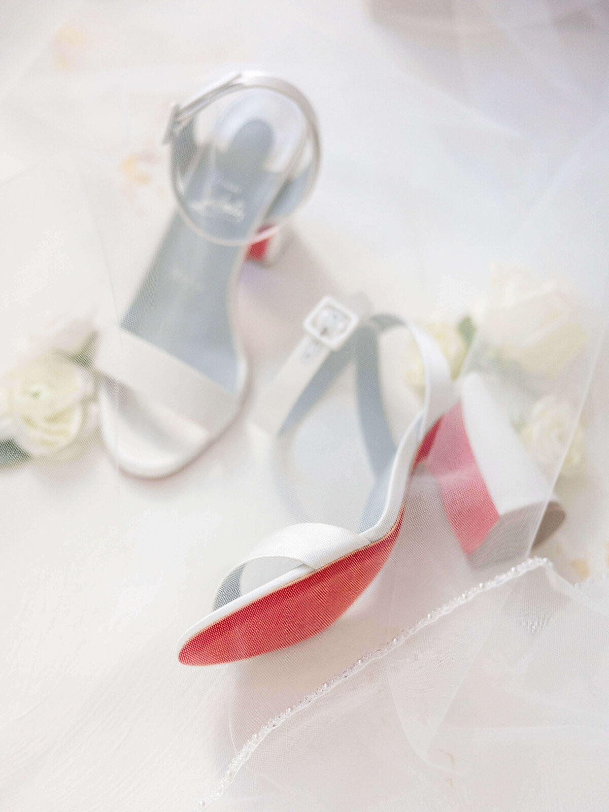 A pair of elegant high-heeled sandals with white and red soles are placed on a soft, white surface. The shoes are surrounded by delicate white roses and sheer fabric, creating a dreamy, romantic setting.