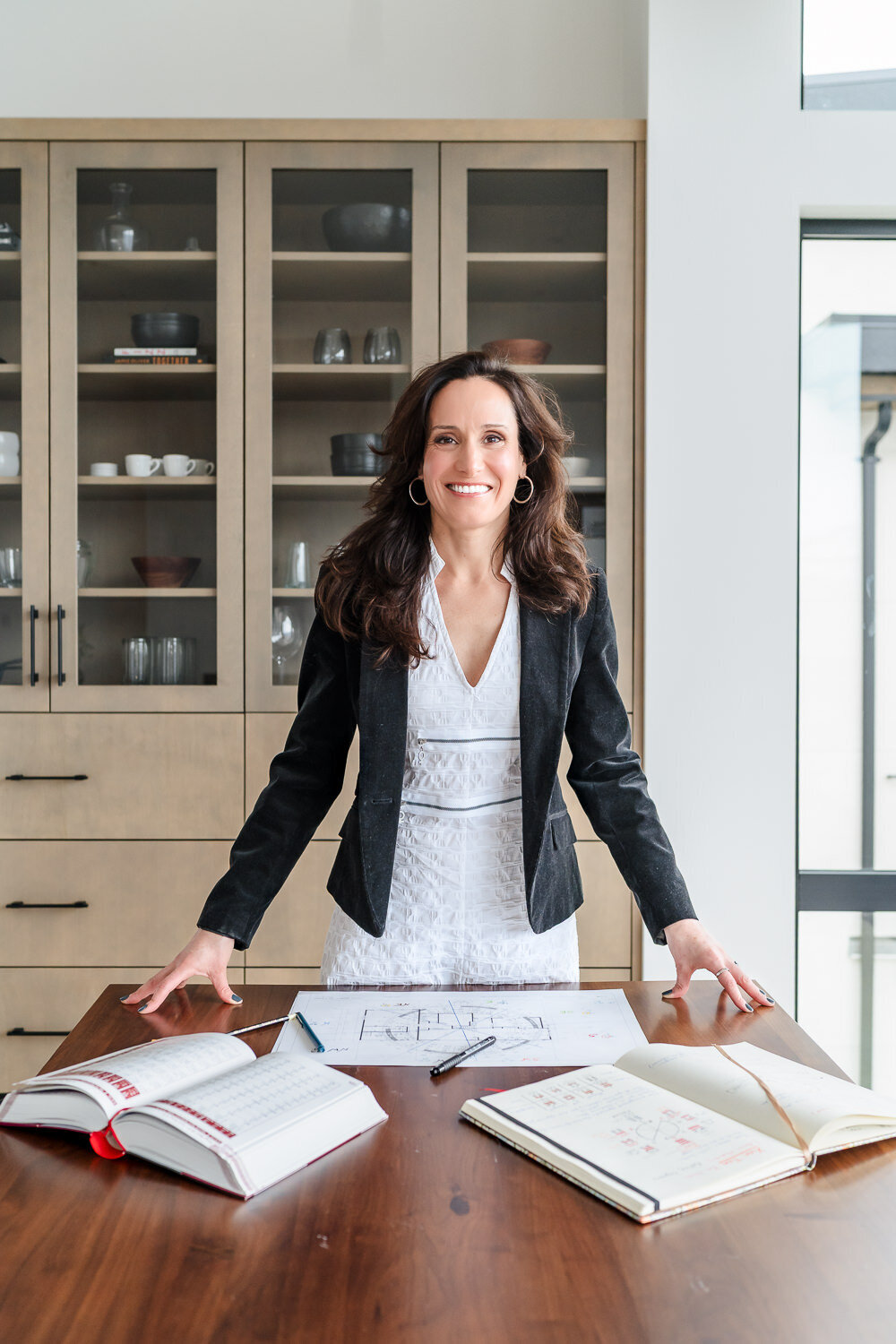 Headshot of business woman leaning forward on table books open in front of her wearing white dress and black blazer