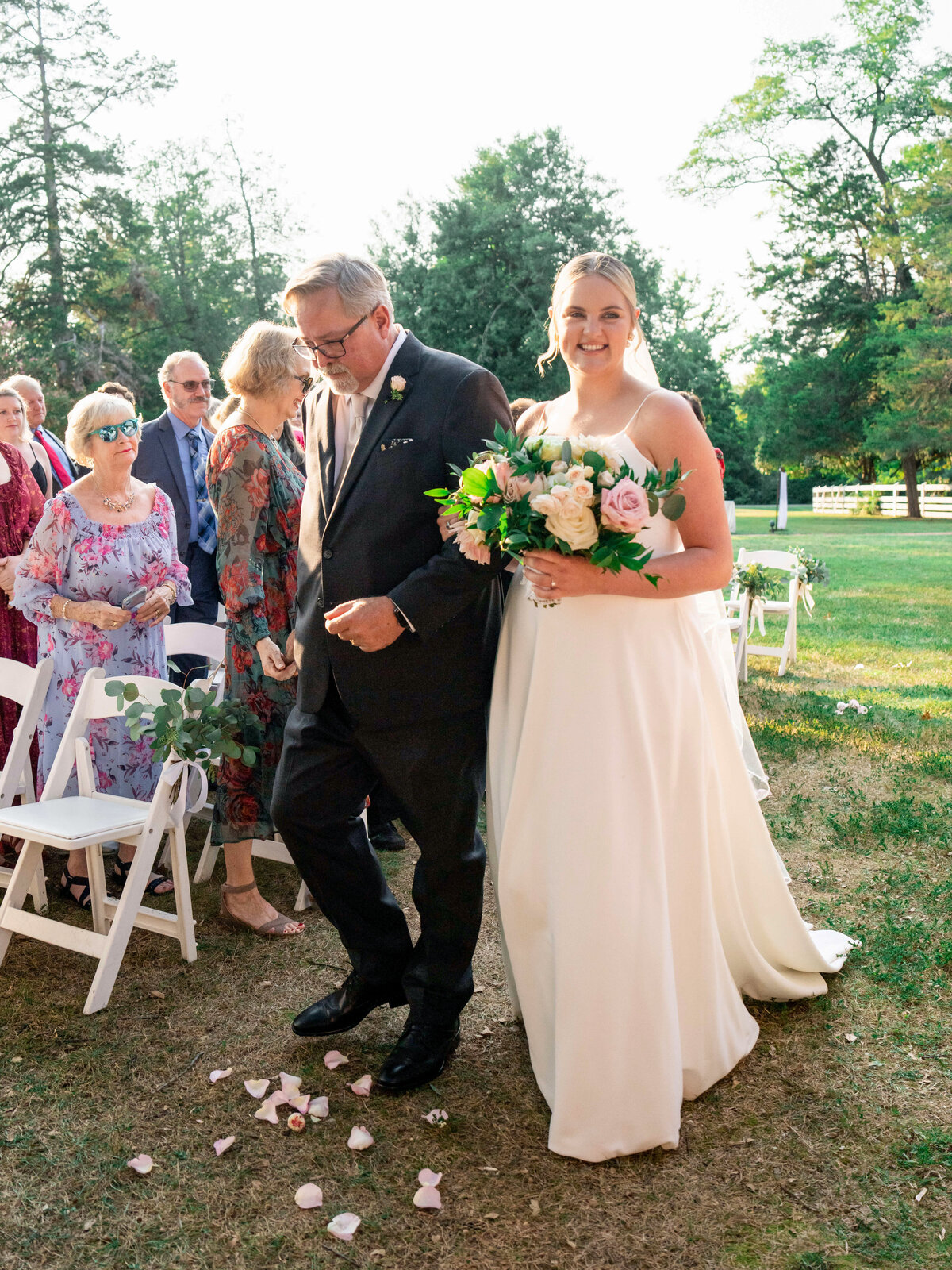A bride in a white dress walks down an outdoor aisle with an older man in a suit. She holds a bouquet of flowers. Guests are seated, watching and smiling. The setting is a sunny, grassy area with trees in the background.