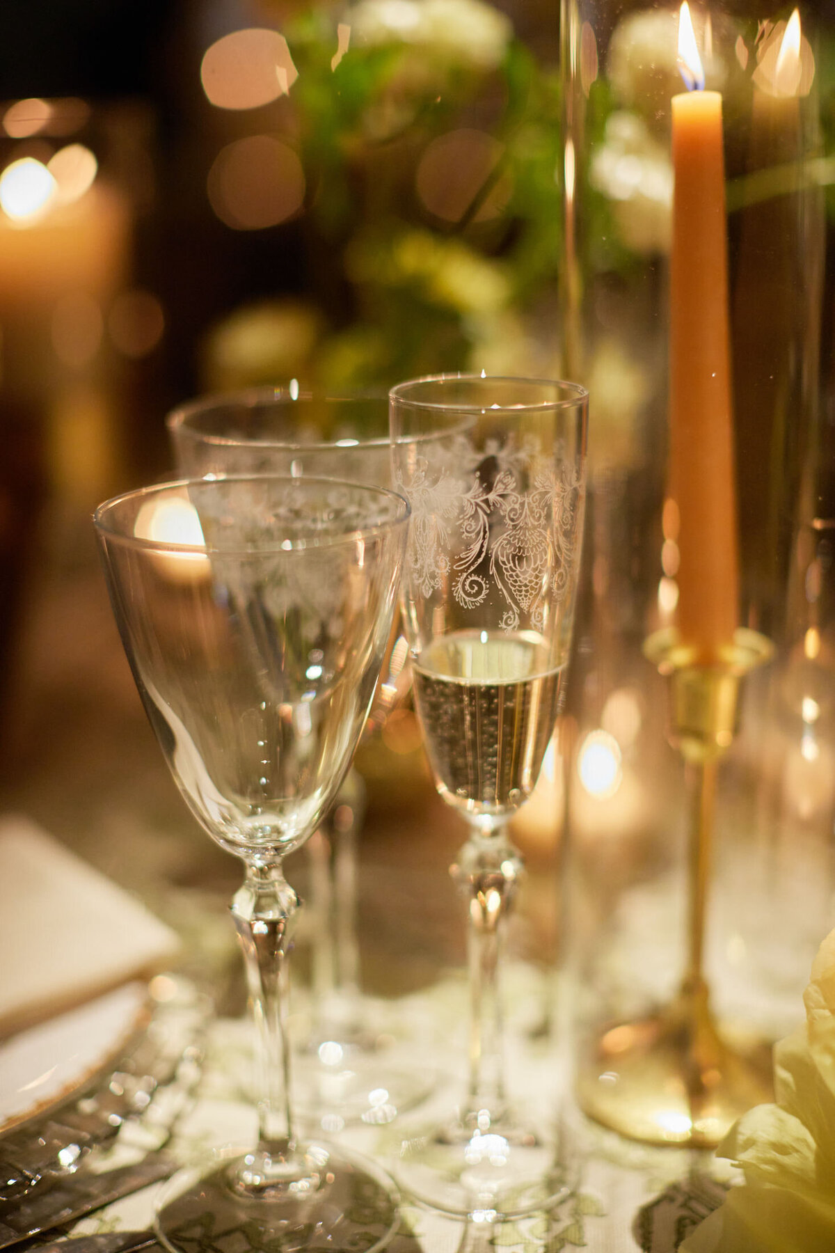 A close-up of an elegant table setting featuring three ornate glasses, one empty and two with champagne. A lit candle in a brass holder stands on the right, with soft-focus greenery and more candles in the background, creating a warm and festive ambiance.