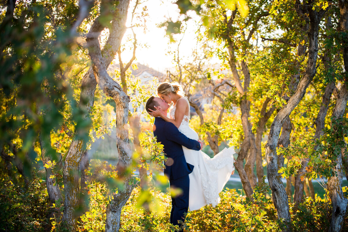 A groom lifts his bride in the air and they share a kiss in the oak trees at The Oaks at Plum Creek.