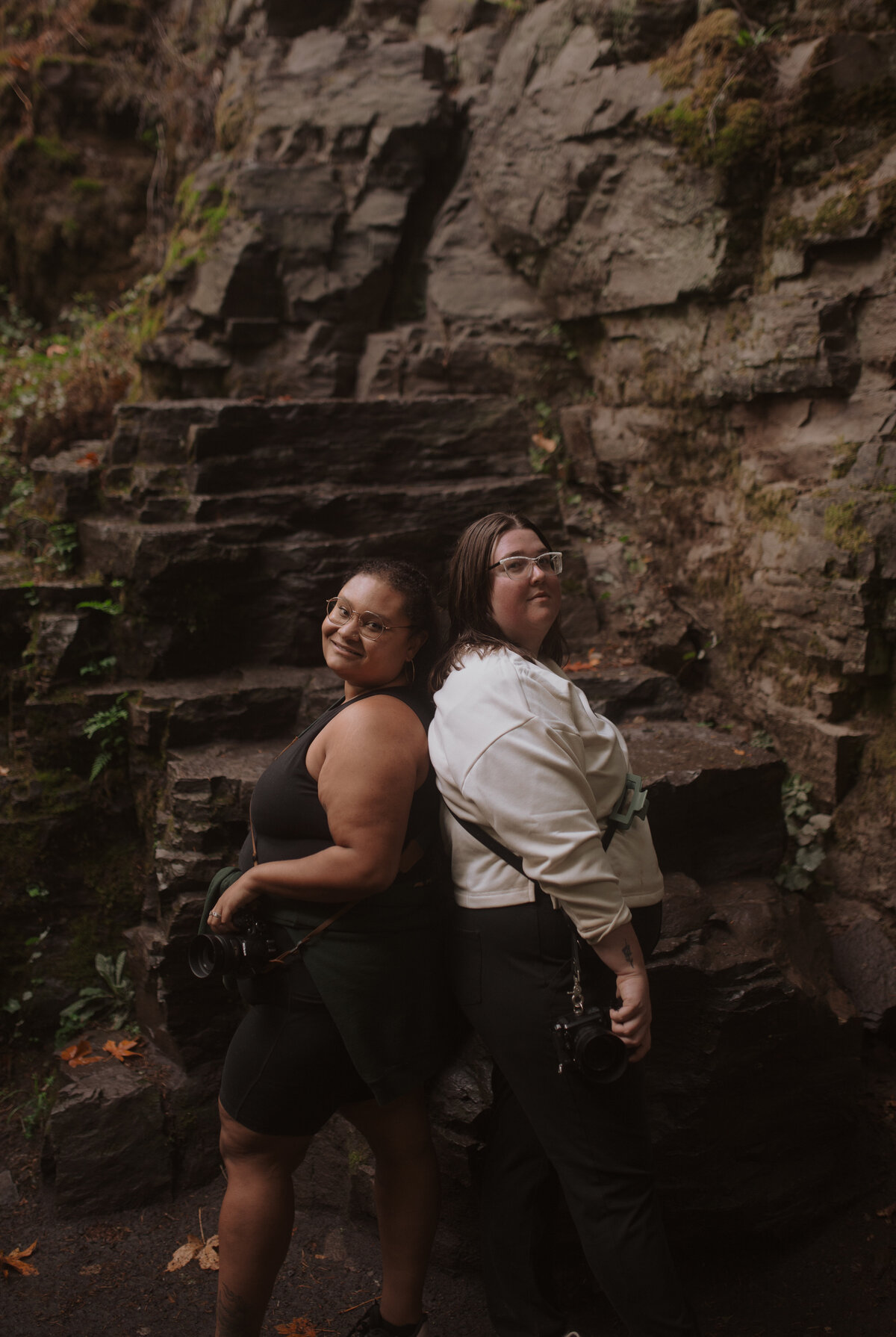 Best friends standing back to back with their cameras on a hike.
