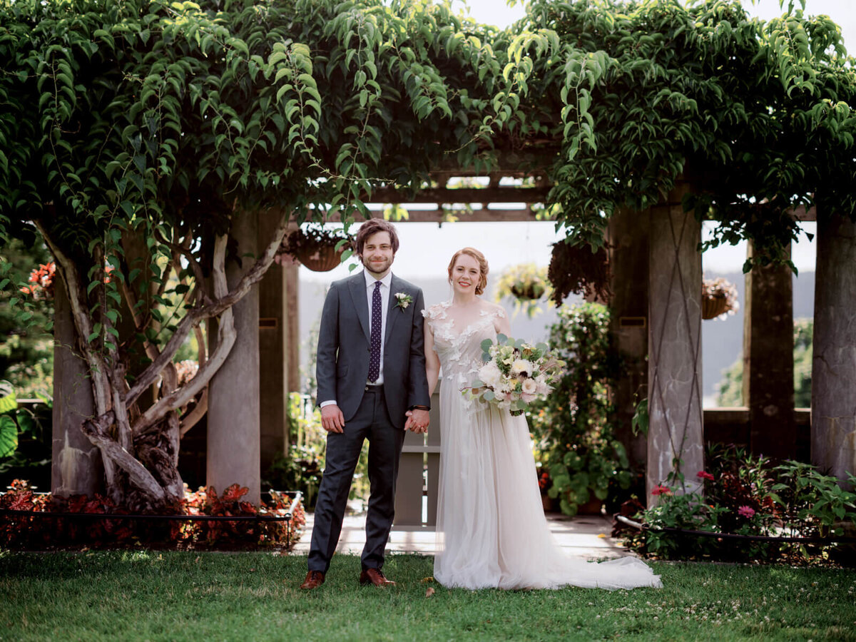 The bride and the groom are standing, on a background of columns with a trellis and thick, green plants on top of the trellis.