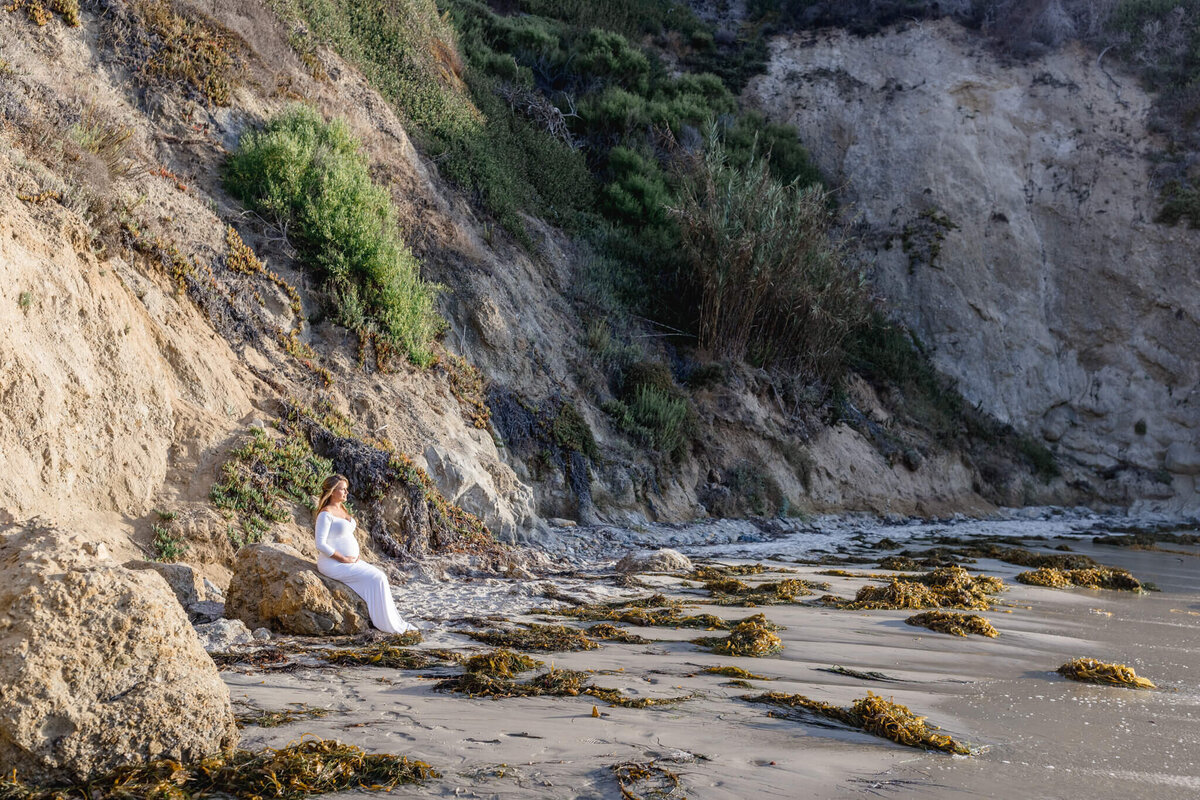 A person in a white dress sits on a rock by a sandy beach, with seaweed scattered along the shore. Steep cliffs covered in greenery rise in the background, under a clear sky.