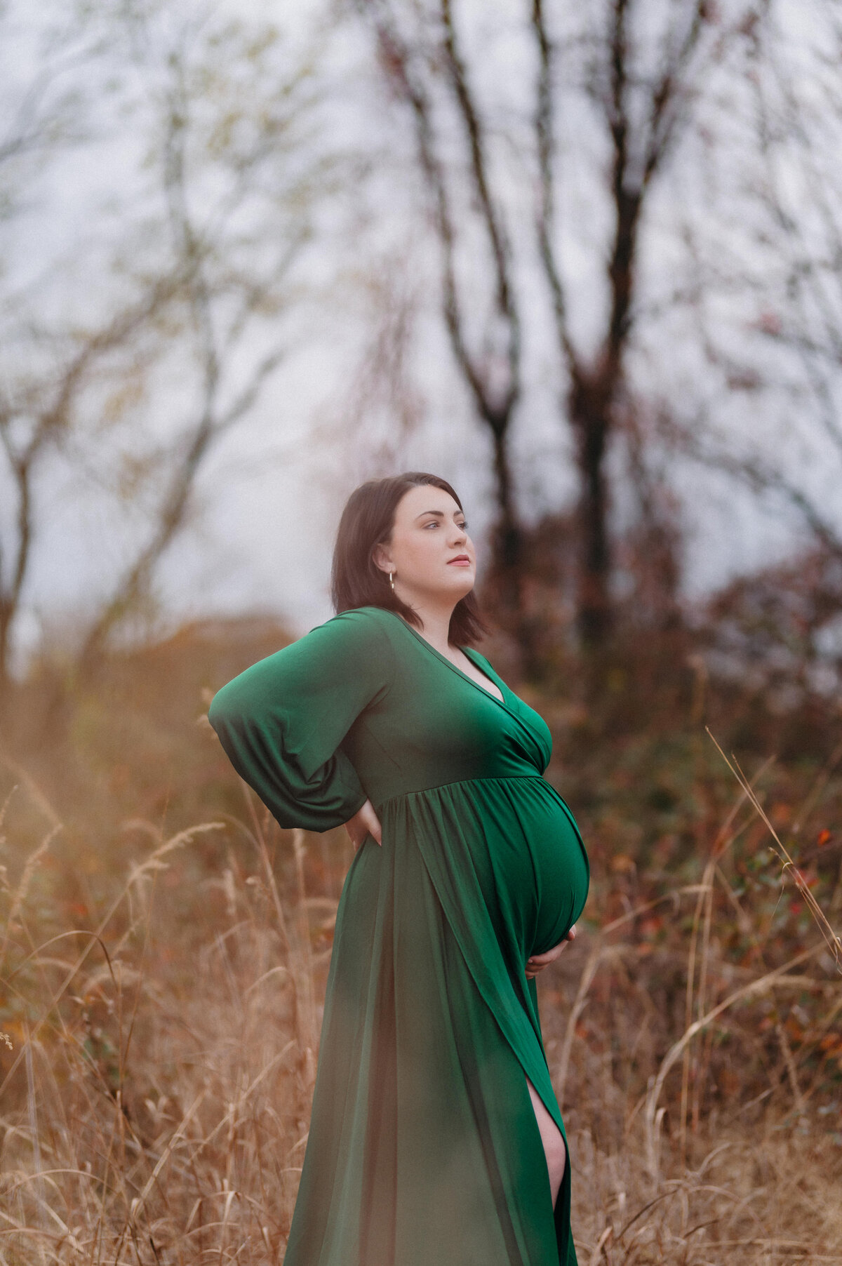 artistic image from low angle of expecting mother in emerald green dress framed by grass in the foreground. location is annapolis maryland.