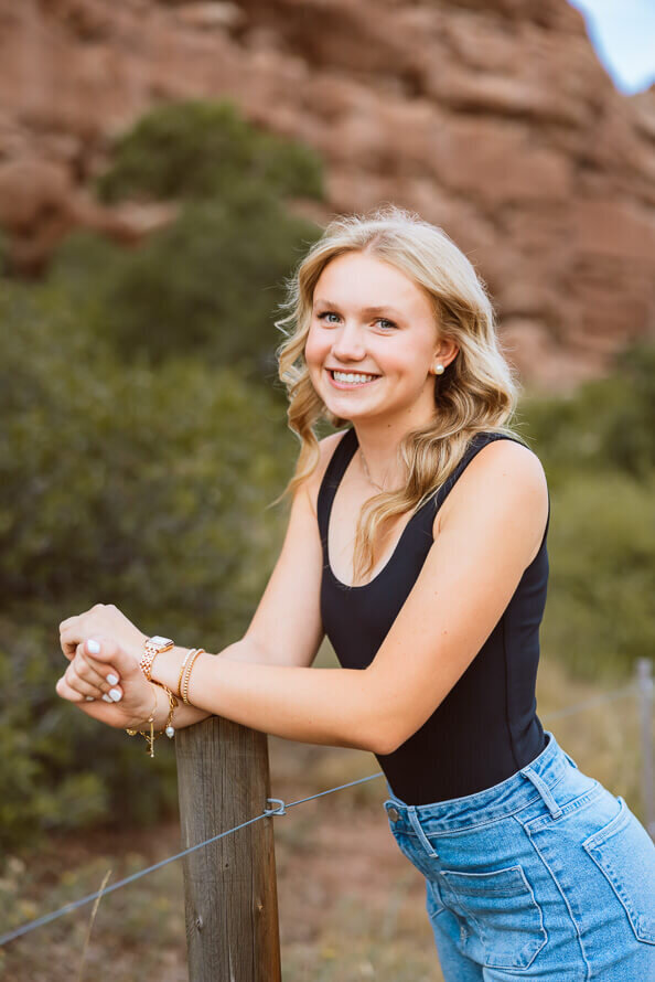 Blond girl in black tank leans on post and smiles with red rocks behind her.