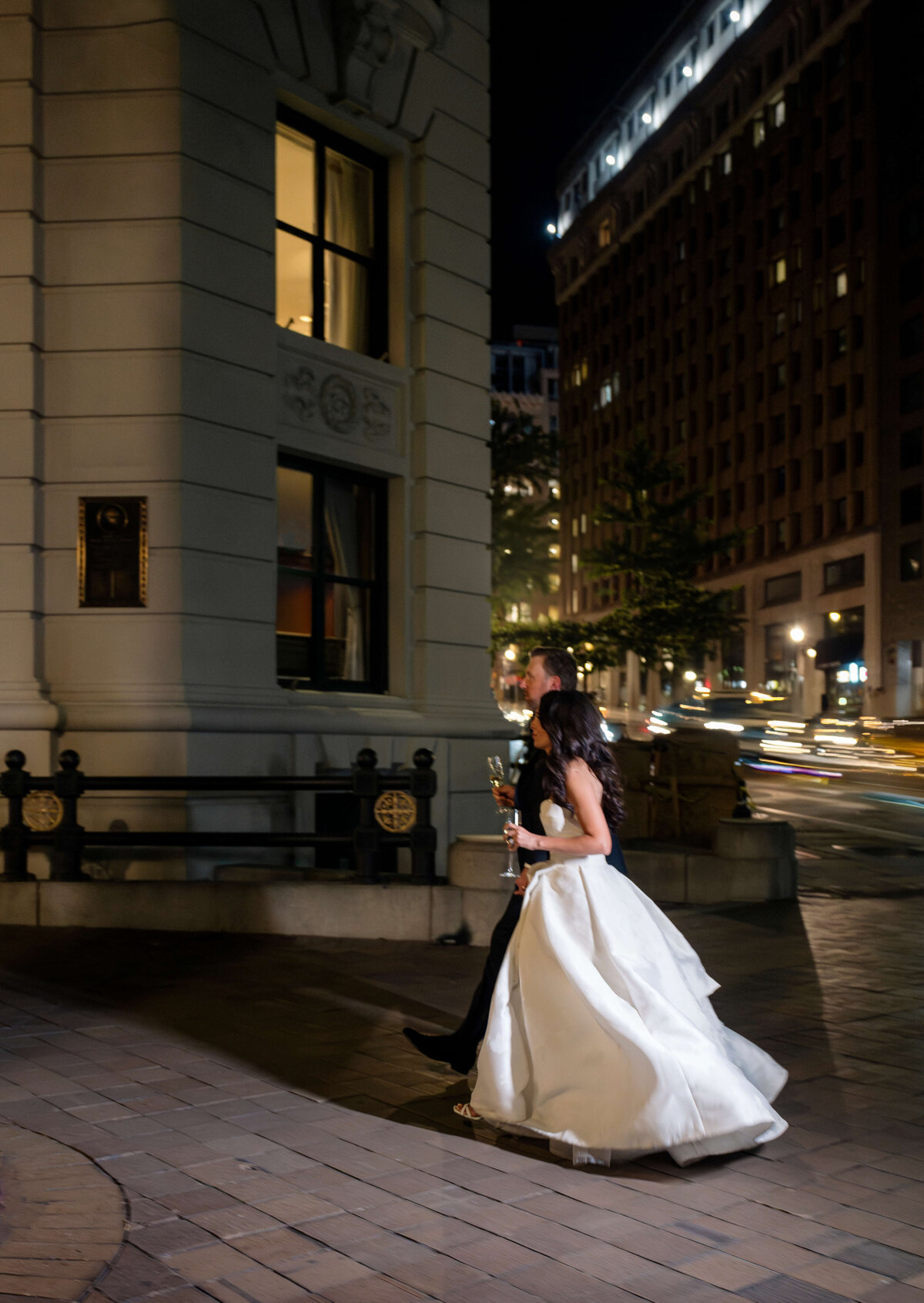 A bride and groom walk along a city street at night, the bride holding her gown and a drink. Tall buildings are illuminated in the background, and the couple appears joyful.
