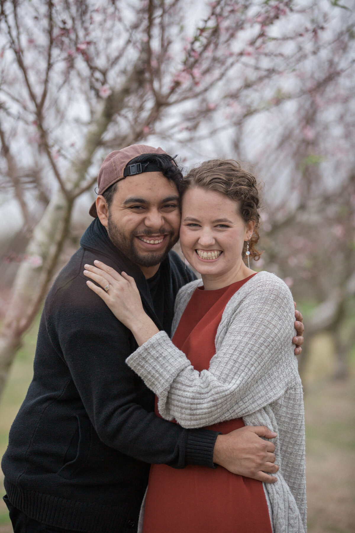 A newly engaged couple smile for the camera in a peach orchard, displaying an opal engagement ring