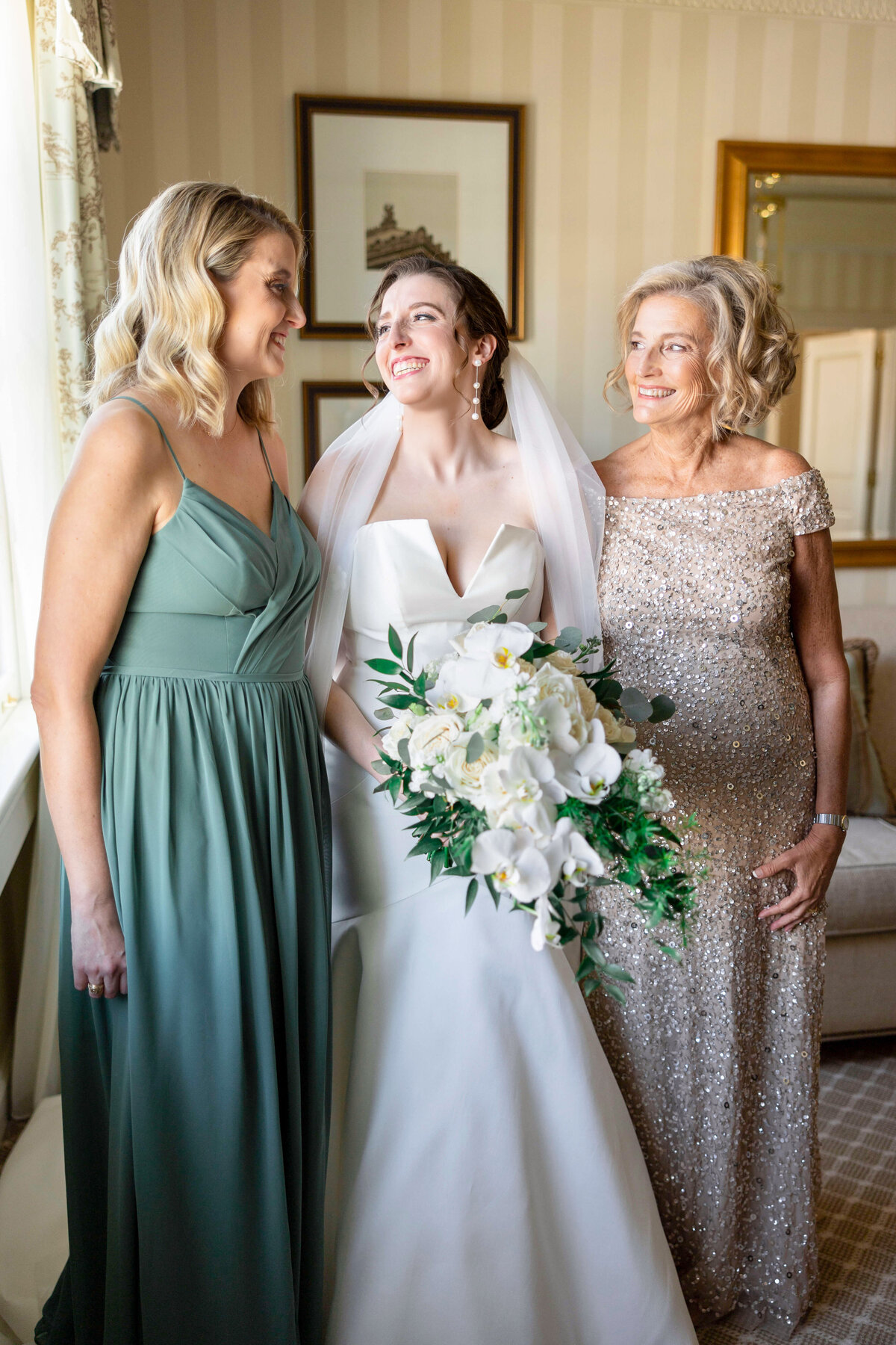 A bride in a white gown holds a bouquet of white flowers while smiling between two women. One wears a green dress, the other a sequined gown. They stand in a room with framed art on the wall and a window with sheer curtains.