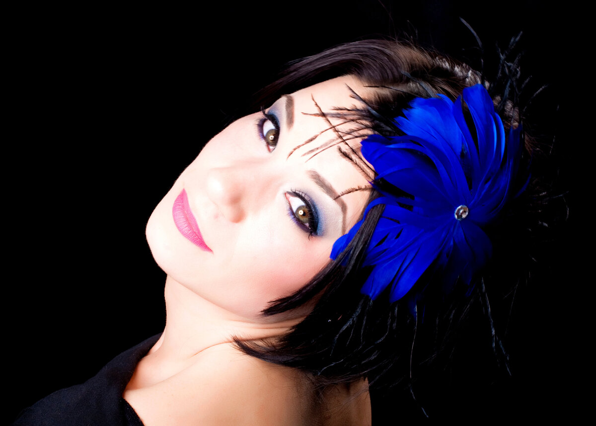 A female performer's close-up headshot of her face wearing a bright blue feather in her hair 