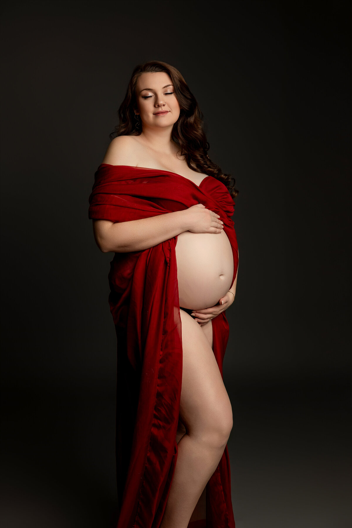 Beautiful expectant mother in a bold red dress, standing gracefully against a dark backdrop, exuding elegance and anticipation for her growing family. Captured by Inland Empire maternity photographer Kaitlyn Dawn Photography.