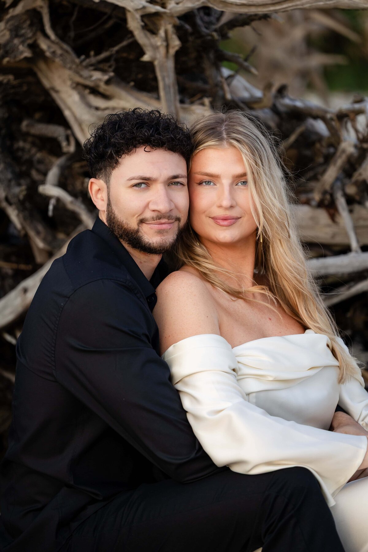 A couple poses closely in front of dramatic driftwood along the shoreline at Driftwood Beach on Jekyll Island. The bride-to-be wears a silky off-the-shoulder white gown, while her fiancé contrasts in a sleek black shirt. Their confident, relaxed expressions and the natural textures of the driftwood create an editorial yet romantic vibe. Captured by Heartful Films, a Jacksonville engagement photographer and videographer known for blending elegance with earthy, coastal backdrops.
