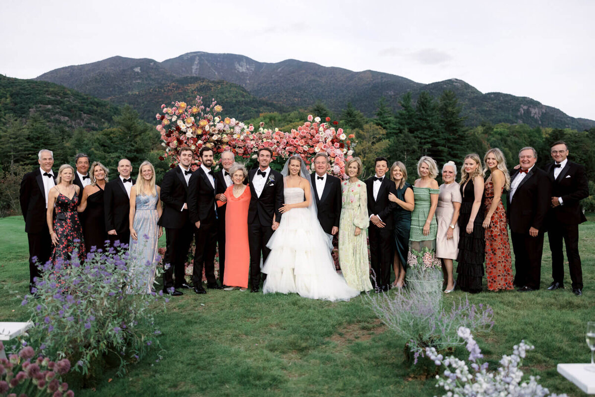 The bride and the groom are with family and friends, with an arch full of flowers and mountains in the background at The Ausable Club, NY.