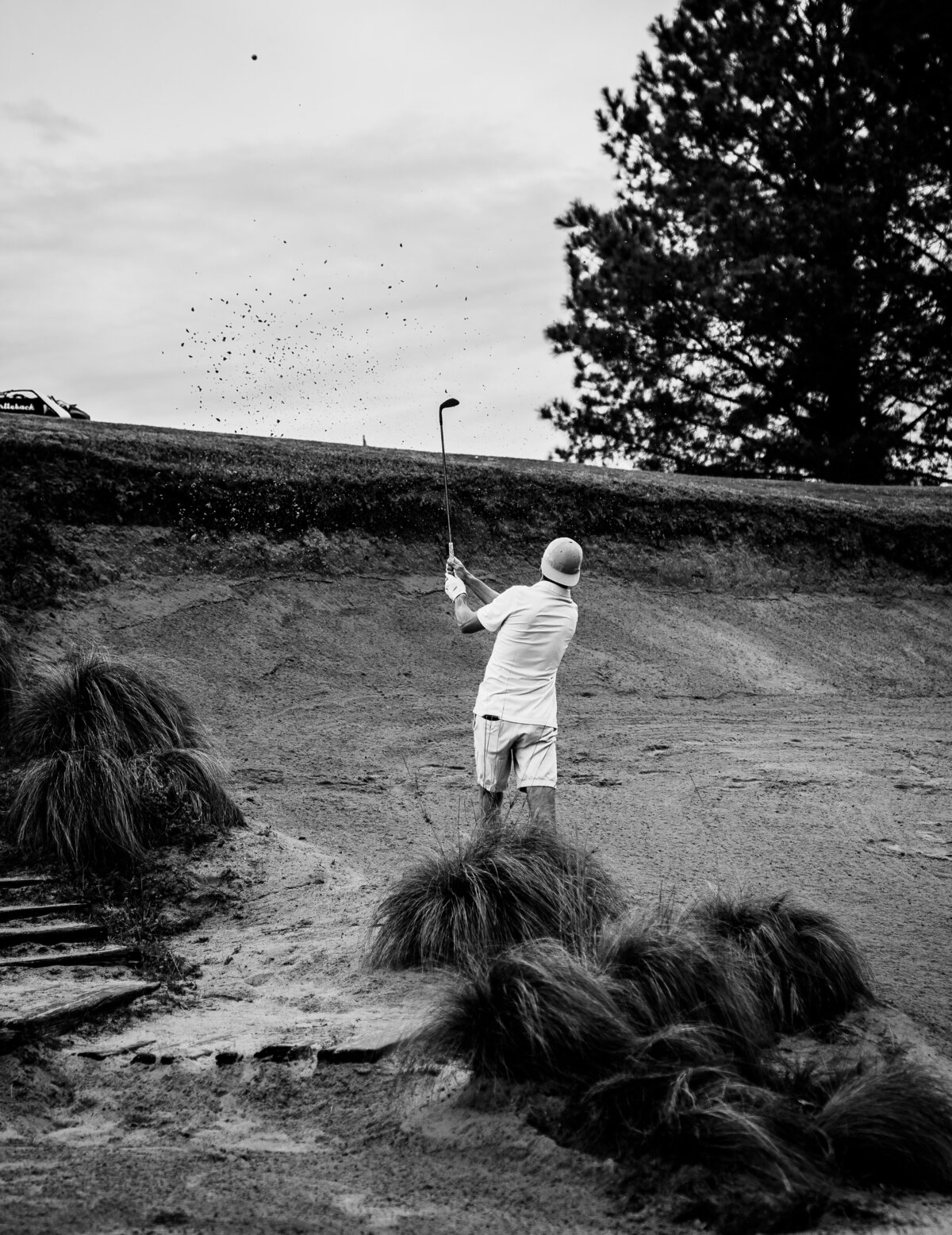 Black and White image of golfer wearing all white with backwards hat hitting ball out of high sand trap at Tobacco Road Sanford, NC. Taken March 2020. 