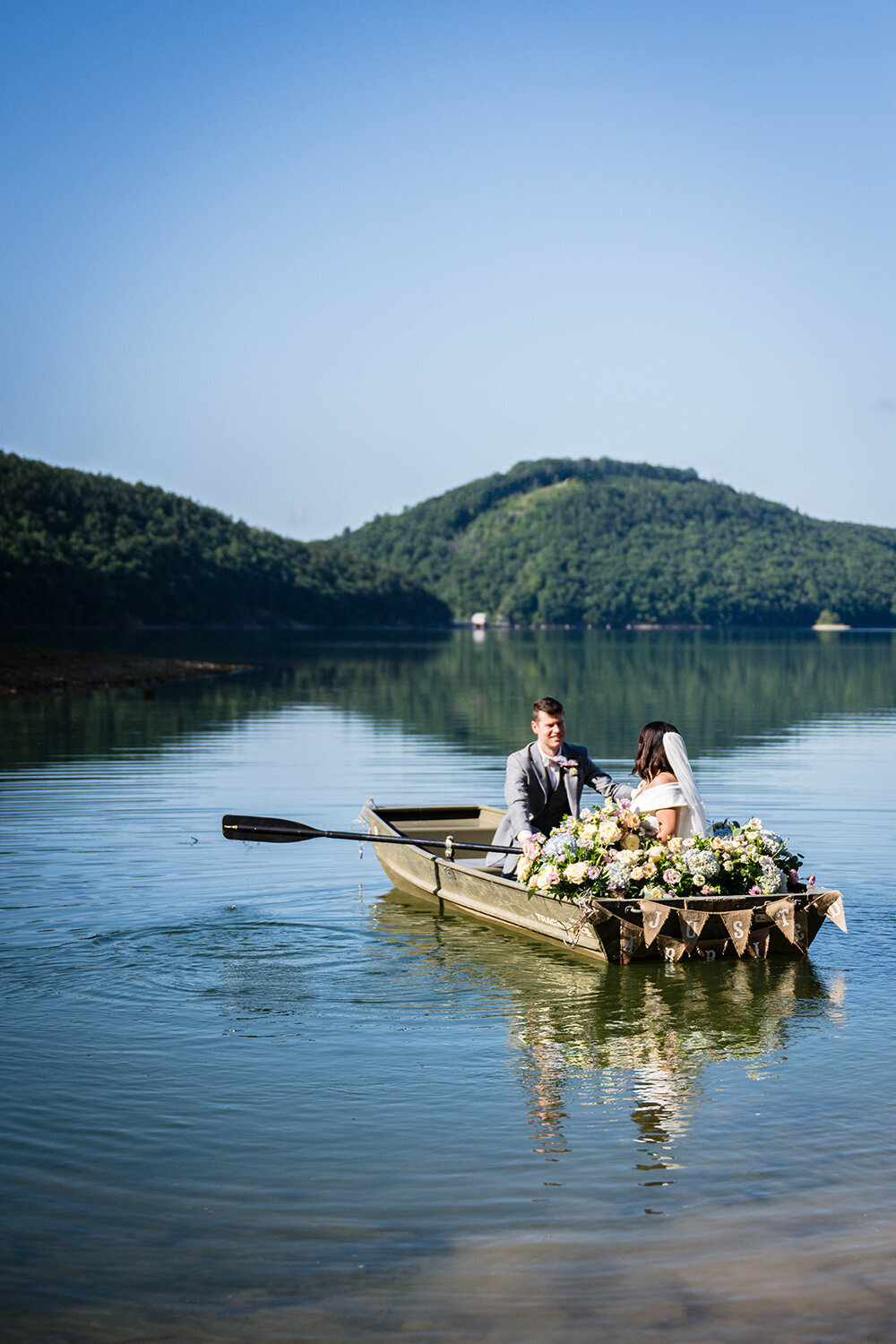 A wedding couple sit in a rowboat in the waters of Carvin’s Cove with the rolling hills of Southwest Virginia in the background. The boat is adorned with pastel flowers and a sign that reads “just married” on the back of the boat. One marrier is rowing the oars while the other sits and looks at him.