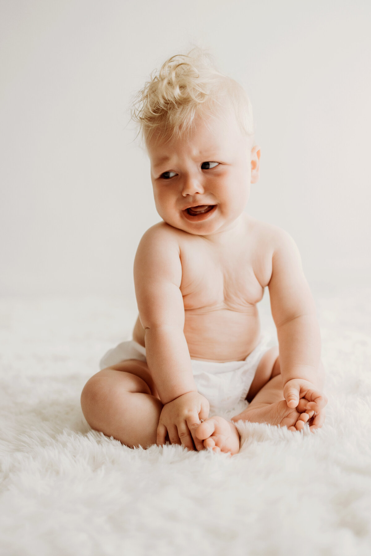 A pouty six month old boy sits up nice and tall on a white rug in a white diaper cover for his milestone photos in Denver. 