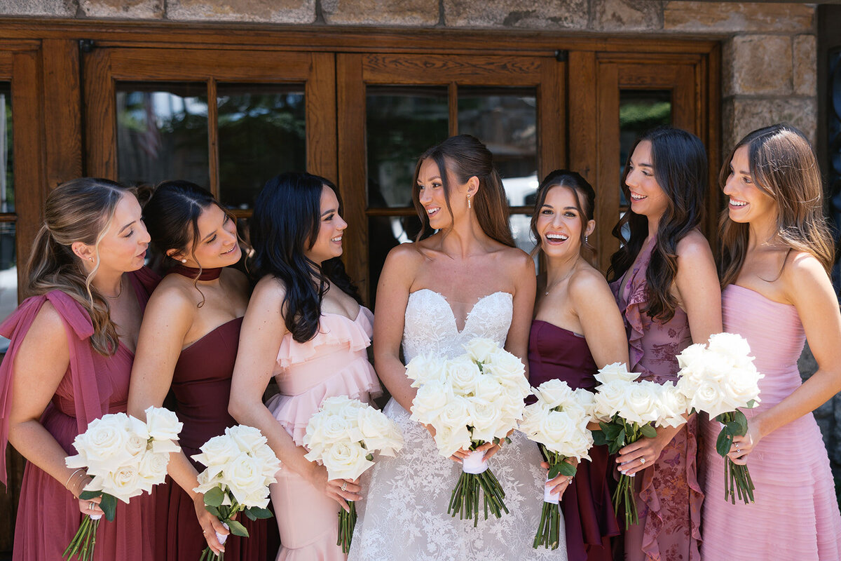 Bridesmaids in mixed pink and burgundy dresses laughing with the bride holding white rose bouquets at Old Edwards Inn in Highlands, North Carolina.