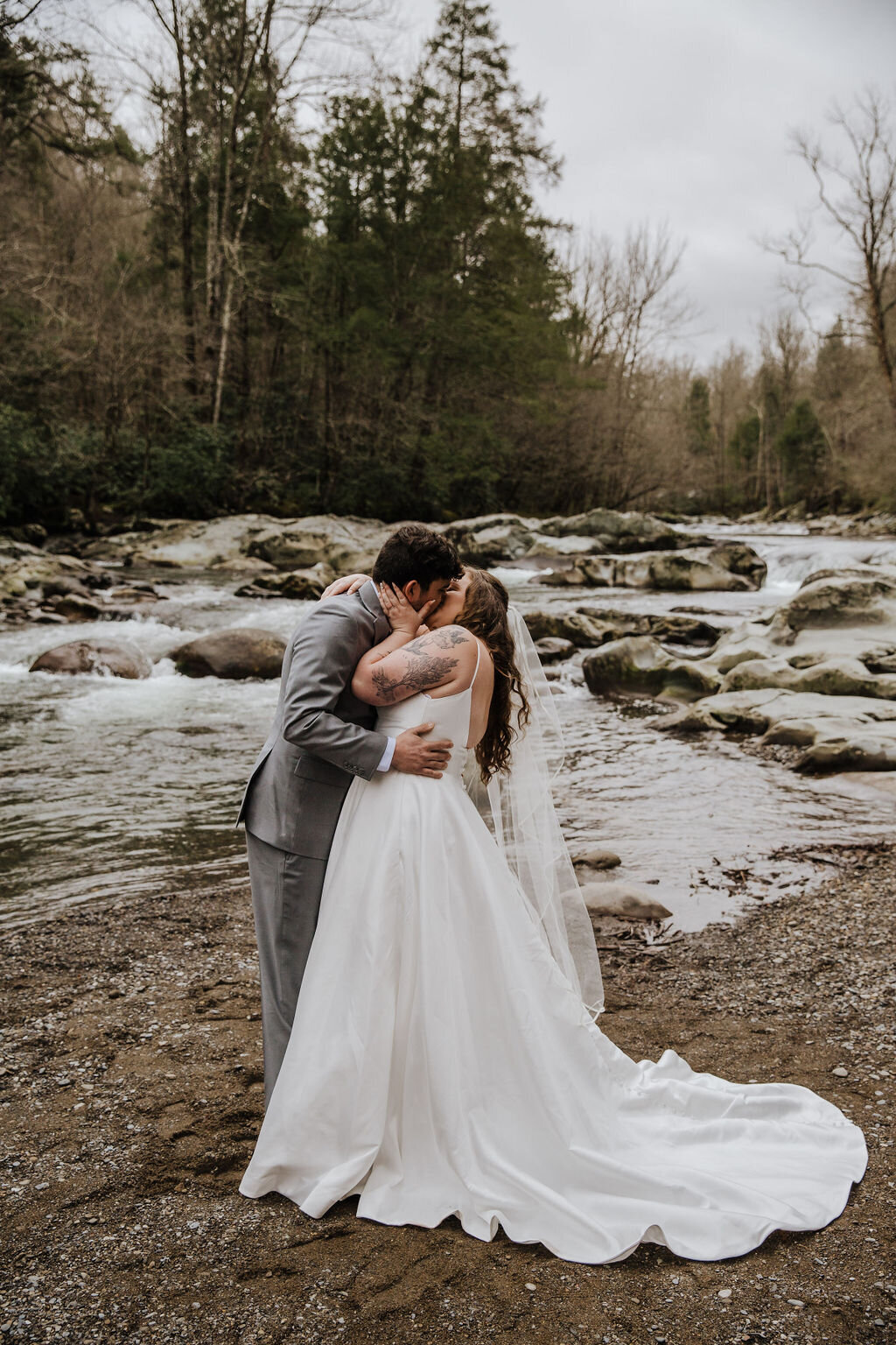 Bride and groom sharing a kiss by the riverside at Greenbrier after eloping to Gatlinburg, with forest trees and smooth rocks surrounding the flowing water.
