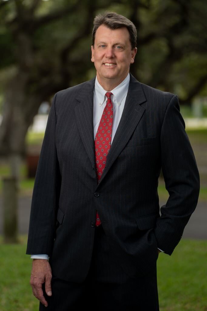 A-man-in-a-dark-suit-and-red-tie-smiles-while-standing-outdoors