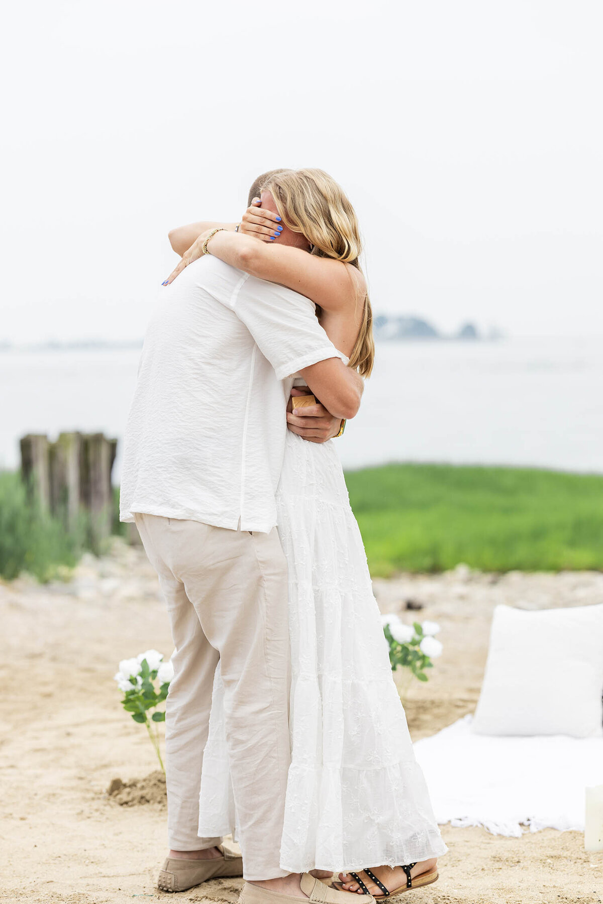 A man wearing a cream white shirt and tan pants is hugging a woman who is wearing a whit dress during a Romantic Coastal Connecticut proposal session 