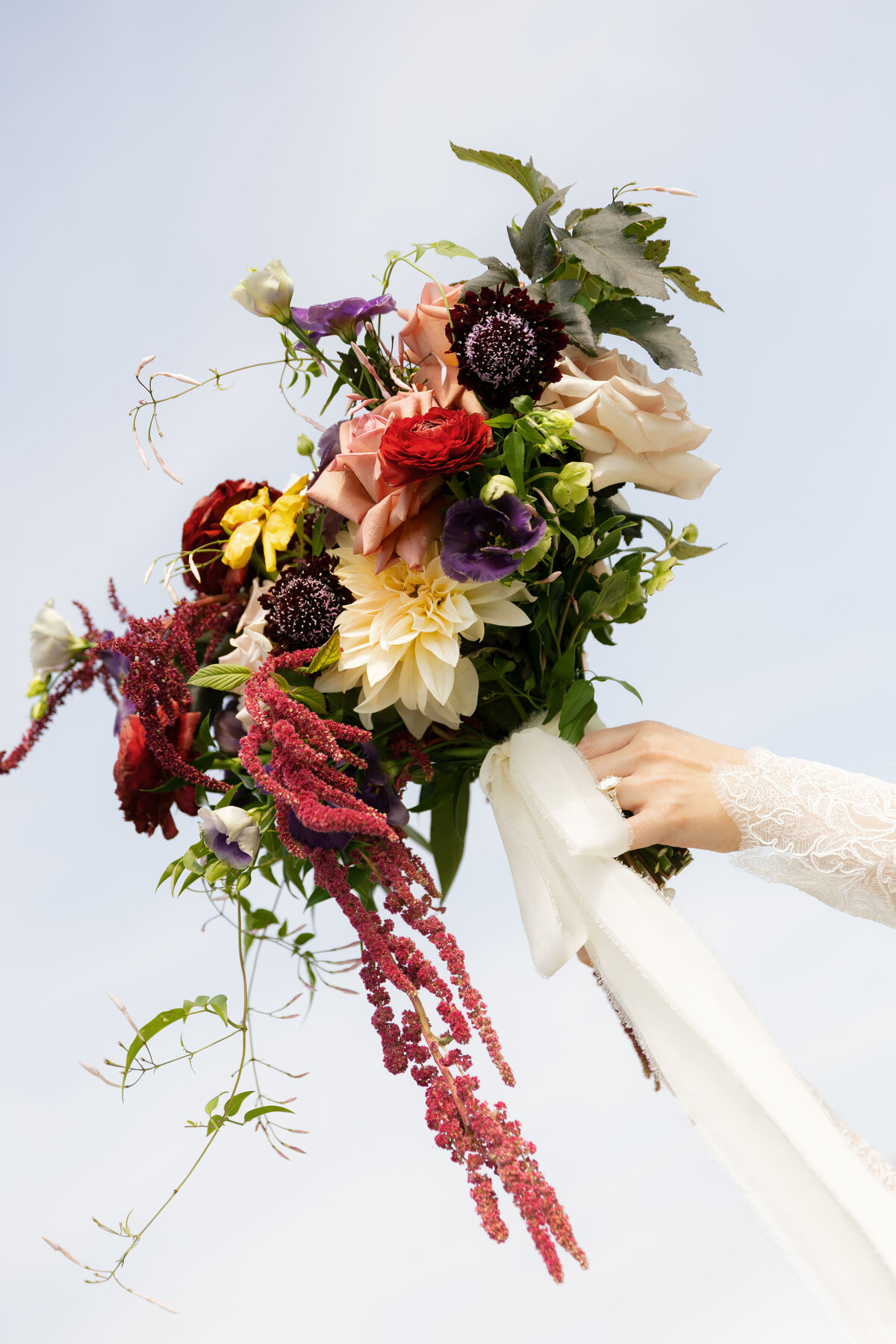 A vibrant wedding bouquet held against a bright sky, showcasing cascading burgundy amaranthus, peach and cream roses, yellow dahlias, purple lisianthus, and trailing greenery. The bride’s lace sleeve and diamond ring are visible as she lifts the bouquet upward.