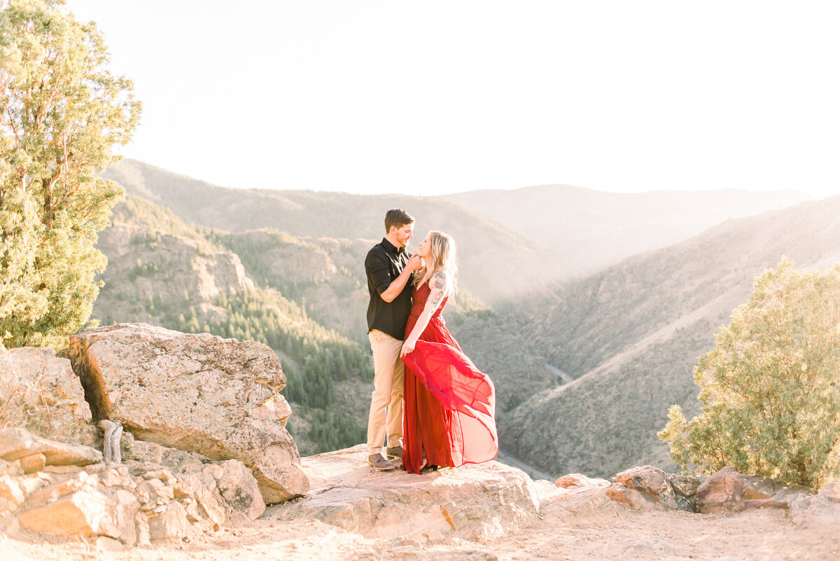 engaged couple in golden colorado posing on the side of a cliff as her red dress flows in the wind and the finace hold her face closely as they  come in for a kiss during a sunset that makes the epic mountain view glow for a must have location for colroado engagement photos by Colorado wedding photographer kari joy photography