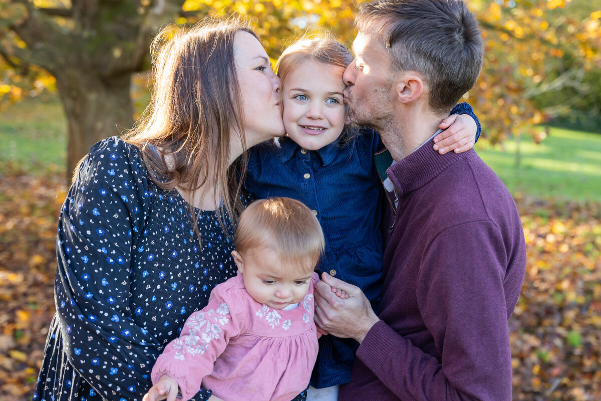 A family of four outdoors in autumn: mother kissing the cheek of a smiling young girl, father kissing the other cheek, while holding a toddler. vibrant fall leaves in the background.