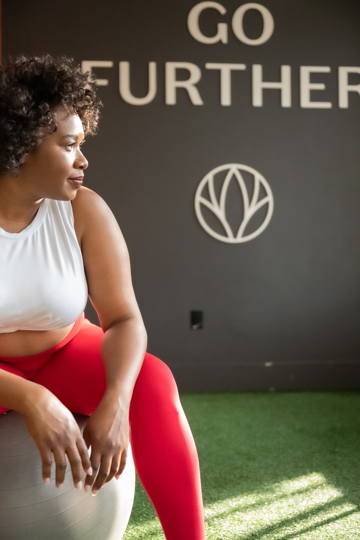 A woman wearing gym clothes, red leggins and white top sitting on a ball smiling and looking at the light on the windw
