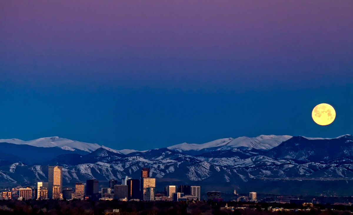 A city skyline at twilight with a full moon rising above snow-capped mountains, reflecting the transition from urban to natural landscapes.