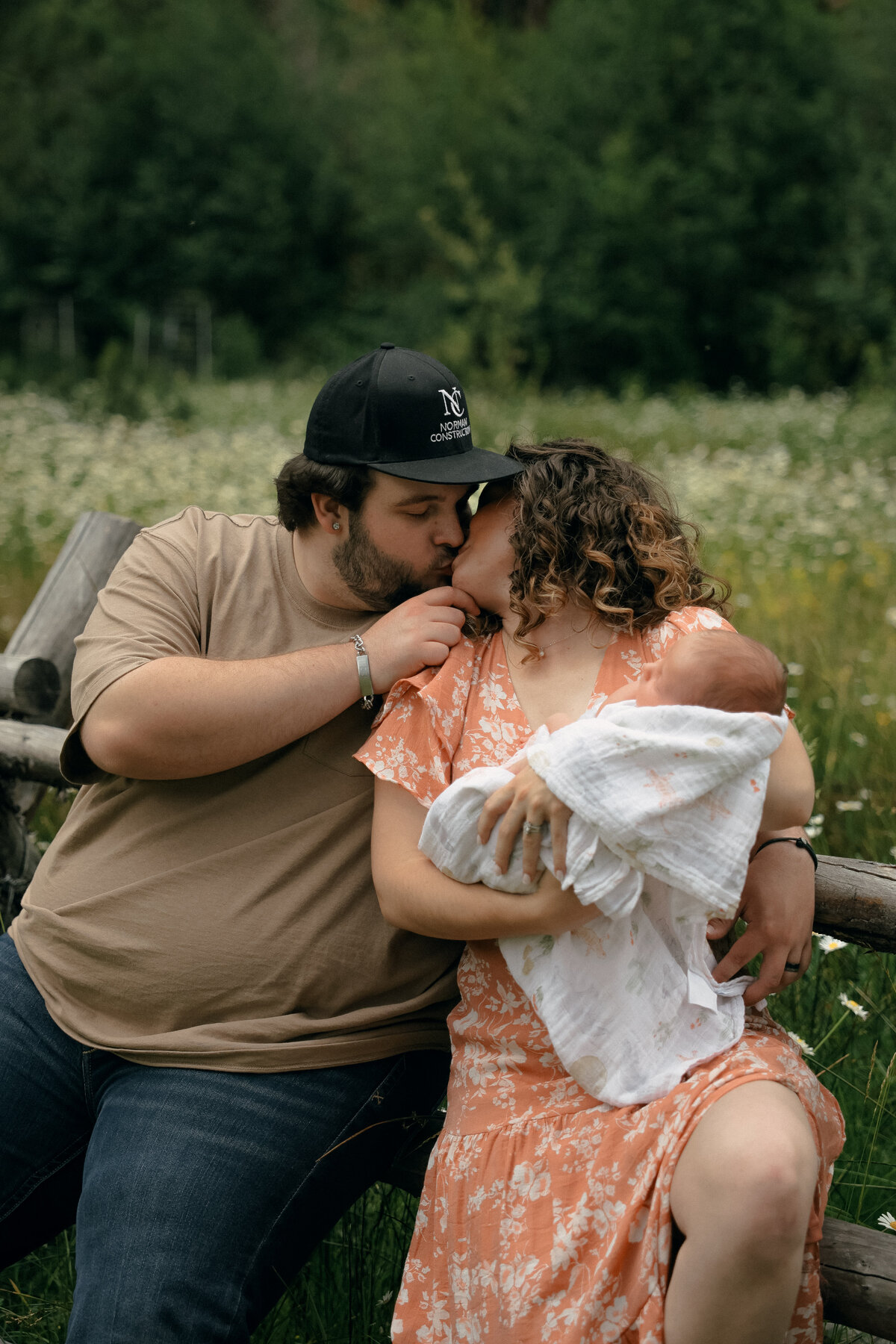 Parents Kissing While Holding Newborn Wrapped in Blanket in Flower Meadow