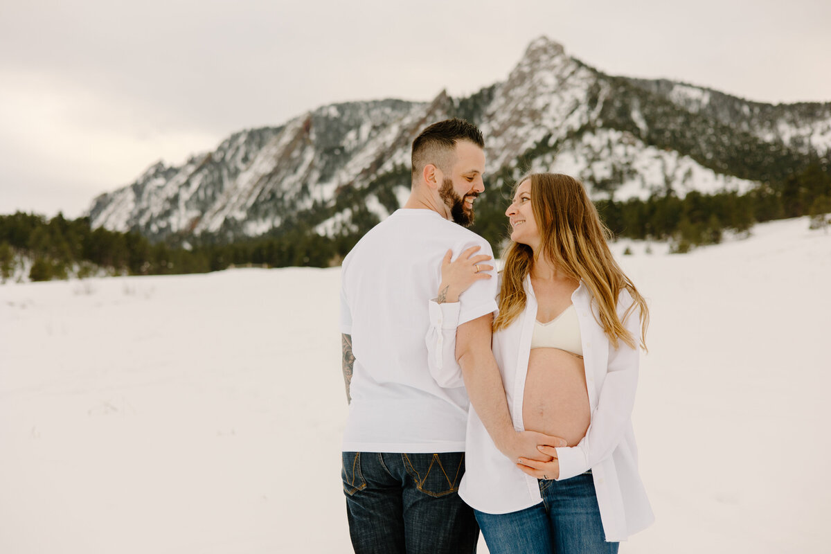A couple poses for a photo during their maternity session in front of the snowy Flatirons at Chautauqua Trailhead.