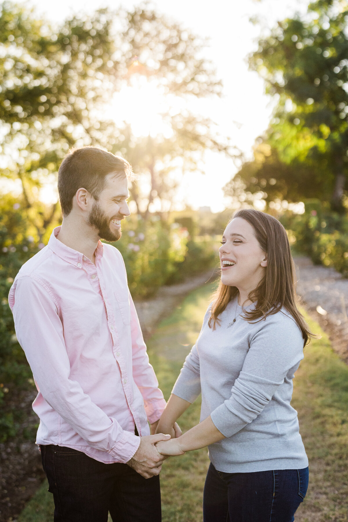 A couple holding hands, smiling, and looking at each other during their engagement session at The Rose Gardens of Farmers Branch in Farmers Branch, Texas. The woman on the right is wearing a grey top and jeans while the man on the left is wearing a salmon dress shirt and jeans. They are backed by many plants and trees.