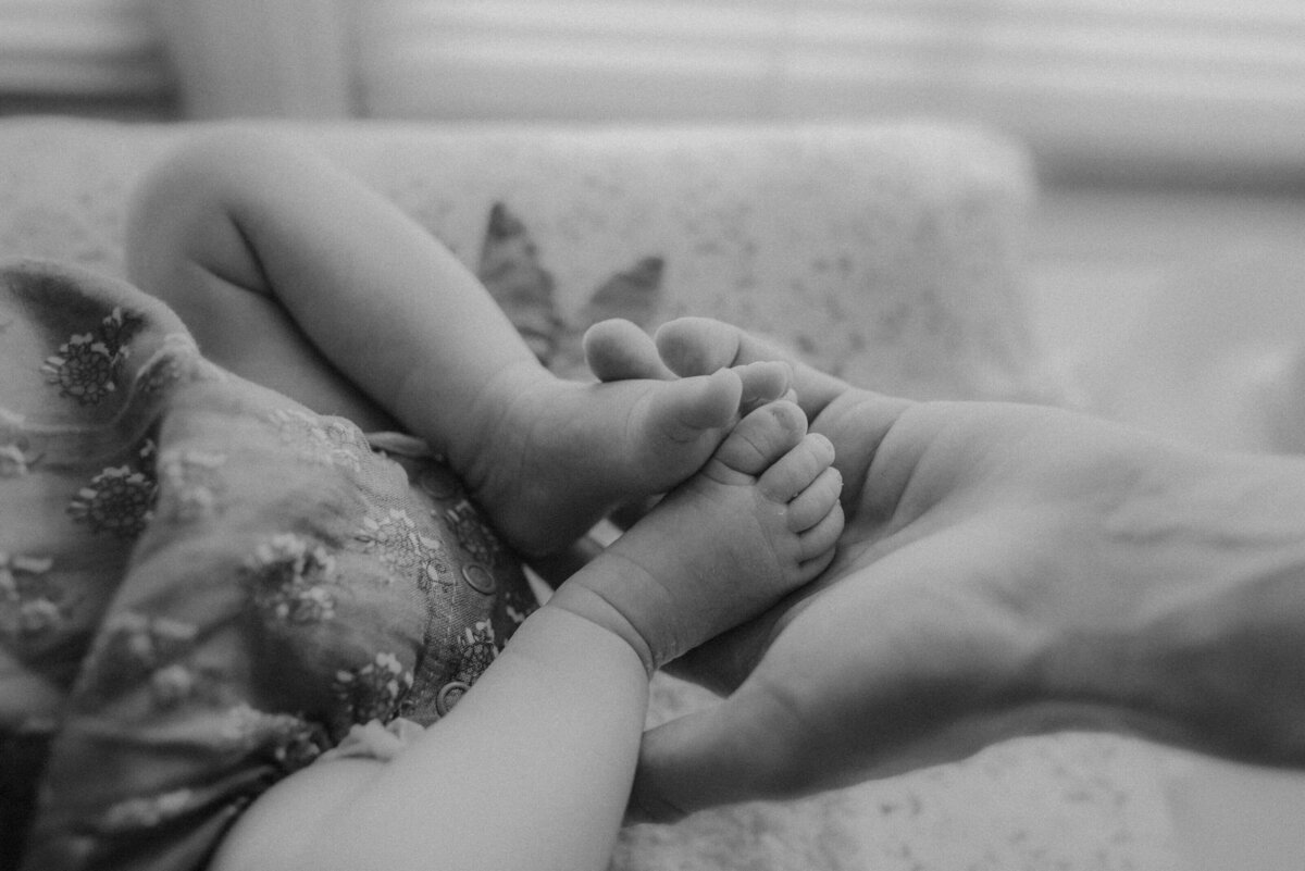 A close-up, black and white image of an adult hand gently holding the crossed feet of a baby lying on a patterned blanket.