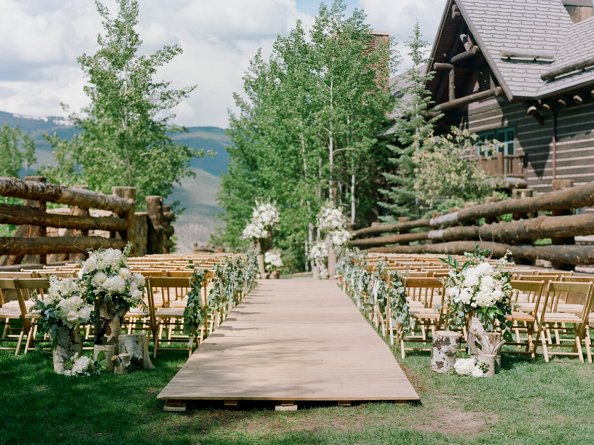 Outdoor empty wedding aisle with wooden runner, and wooden chairs, decorated with white flowers on logs, a wooden lodge is in the background
