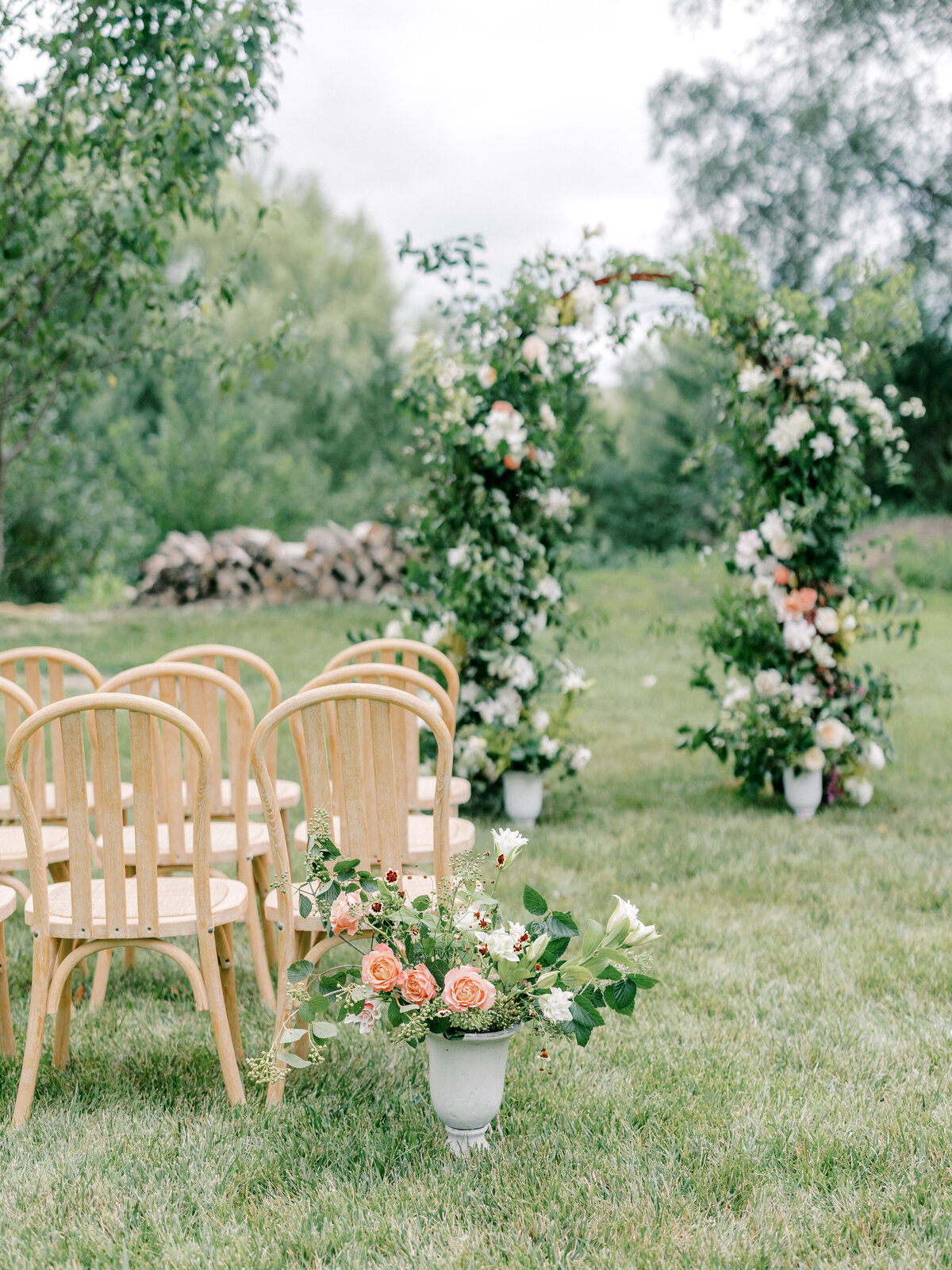 Wedding ceremony decor with peach colored flowers in a vase next to wooden chairs with a greenery floral arch in the background