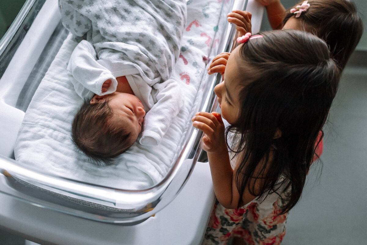 sisters standing over hospital bassinet and looking in to see newborn brother
