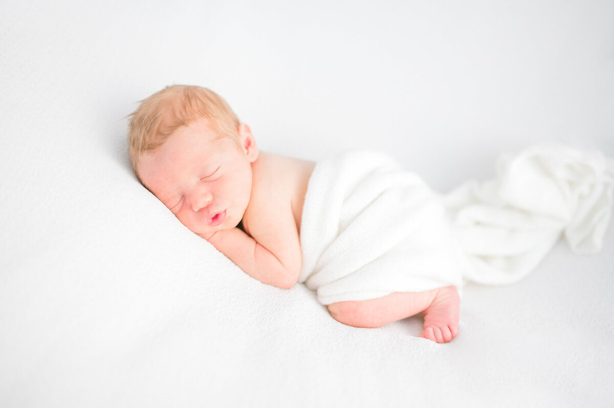 Baby on tummy on white backdrop with white swaddle drapped over