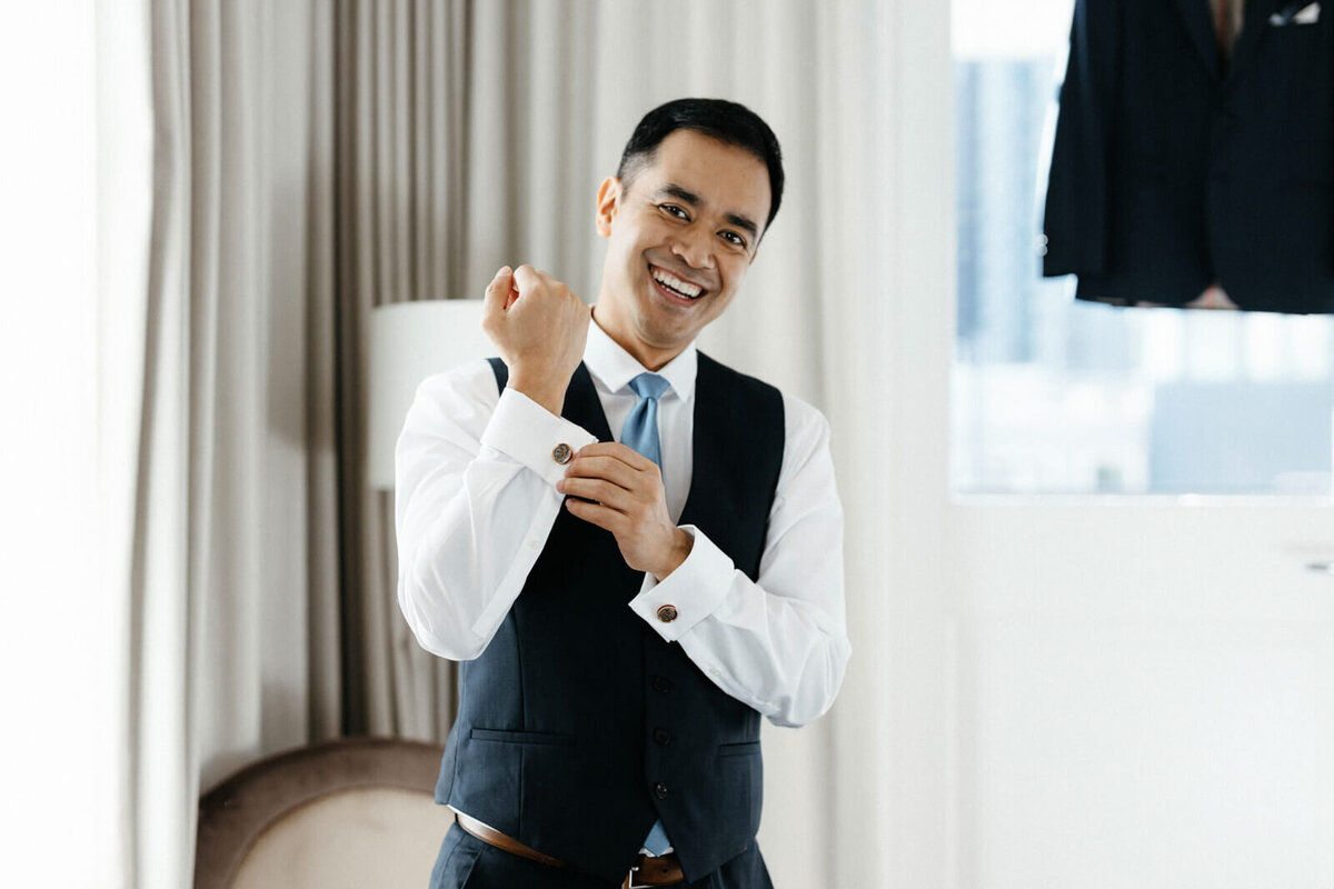 The groom, who is wearing a white long-sleeved polo and a black vest, is smiling while closing the gauntlet button.