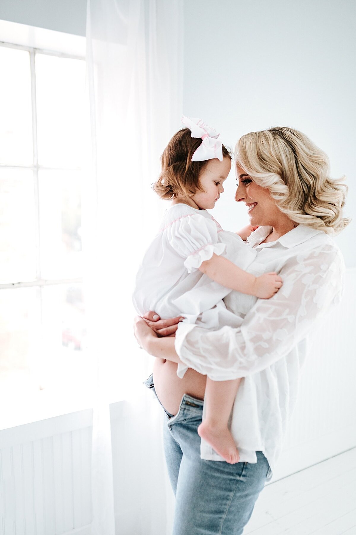 pregnant woman wearing white holding toddler daughter standing in natural light studio