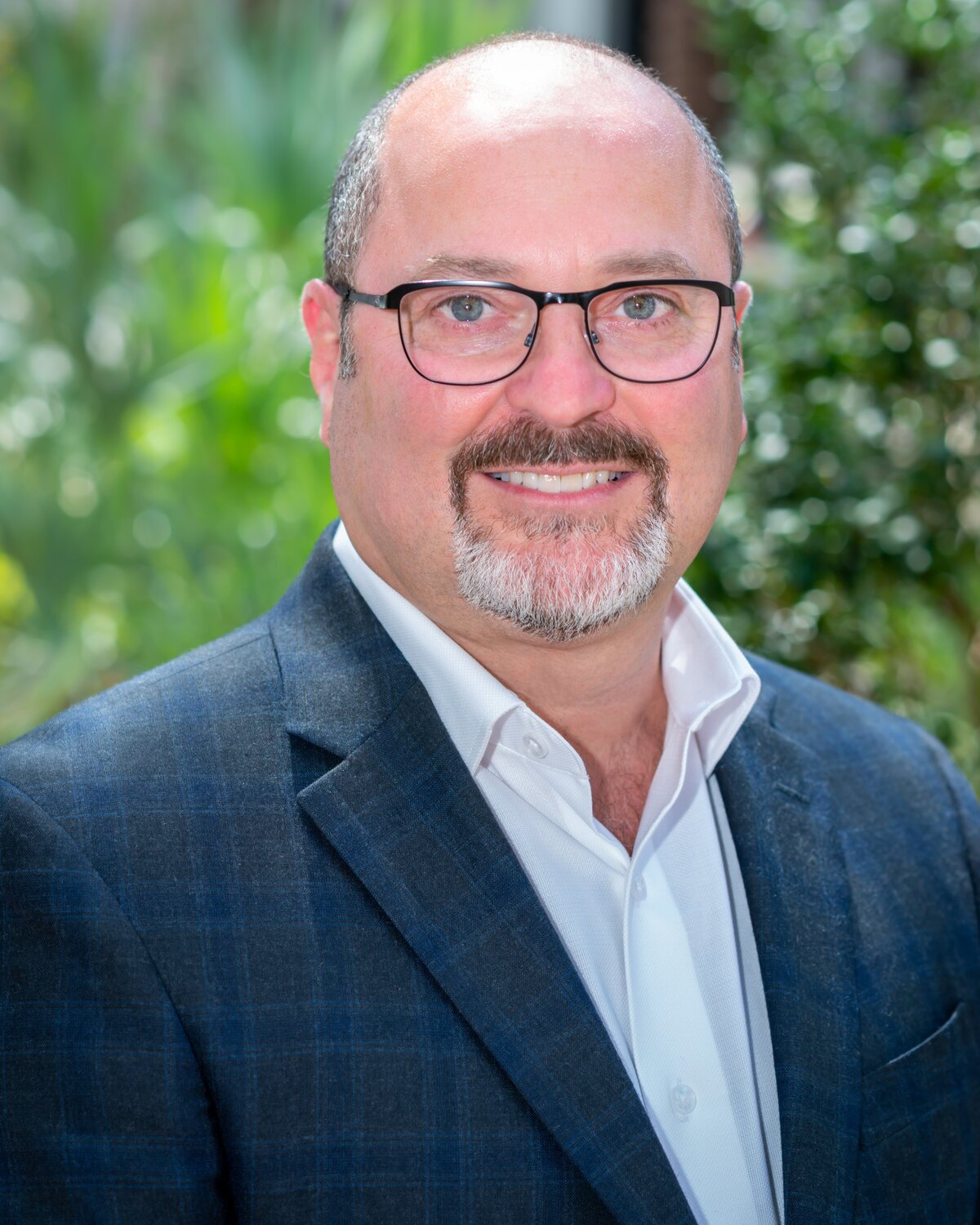 A bald man with glasses poses in a suit in front of green trees for his outdoor headshot session.